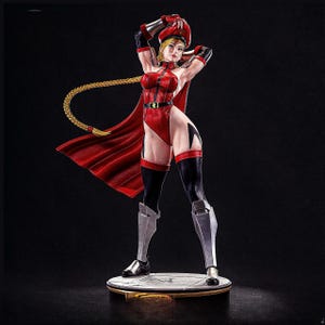 Puede incluir: Figura detallada de un personaje femenino con un body, capa y gorra rojos. La figura tiene cabello rubio en una larga trenza y posa con los brazos levantados. La base es circular con un borde dorado, sobre un fondo oscuro.