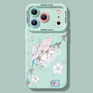 May include: A light mint green phone case featuring a camera lens protector with the text GOOD LUCK. The back displays a graphic of an open box containing apple-shaped glasses, surrounded by white flowers, blue ribbons, and small stars.