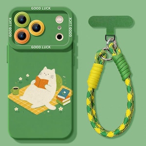Puede incluir: Funda de teléfono verde con una ilustración de un gato leyendo un libro. La funda tiene un protector de cámara con el texto "GOOD LUCK". También se incluye una correa de teléfono verde a juego con cuerda amarilla y verde.