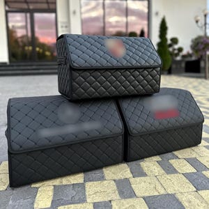 May include: Three black rectangular storage organizers with a quilted diamond pattern on the exterior. Each organizer features a hinged lid and a structured shape, designed for vehicle trunk organization with a sleek, uniform appearance.