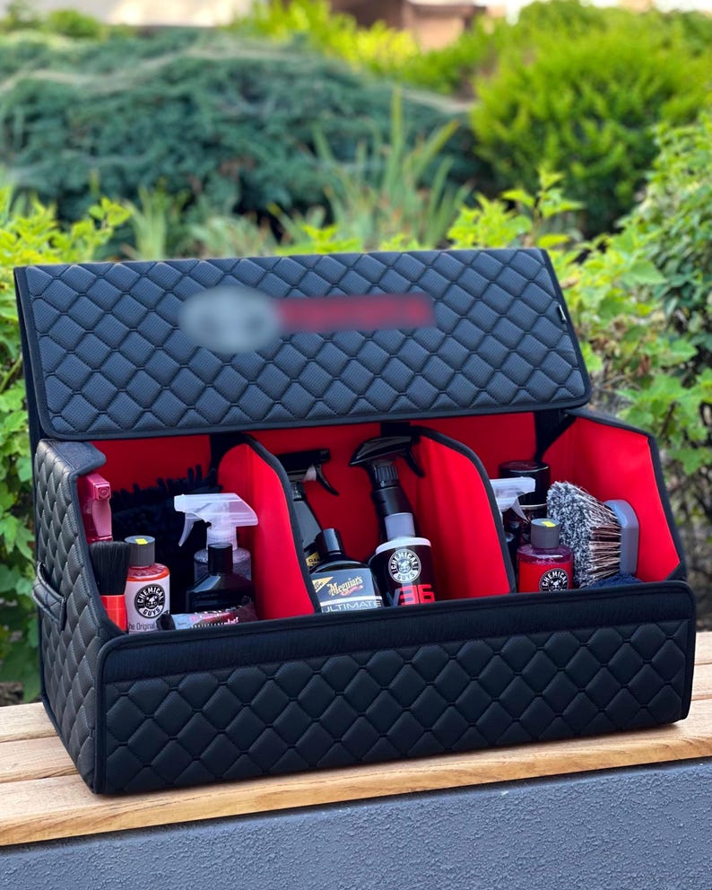 May include: Black quilted car organizer with red interior compartments, holding various car detailing products. The organizer is open, displaying bottles, sprayers, and brushes. The background features green foliage.