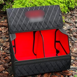 May include: Black car trunk organizer with a quilted pattern on the exterior. The interior is bright red and divided into compartments. The organizer is open, revealing the interior and the dividers.
