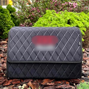 May include: A black, quilted car organizer with a diamond pattern. The organizer has a rectangular shape with a folding lid. The background features greenery and mulch.