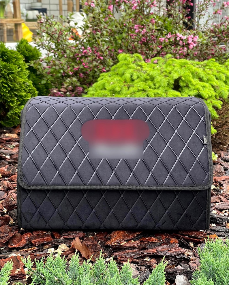 May include: A black, quilted car organizer with a diamond pattern. The organizer has a rectangular shape with a folding lid. The background features greenery and mulch.