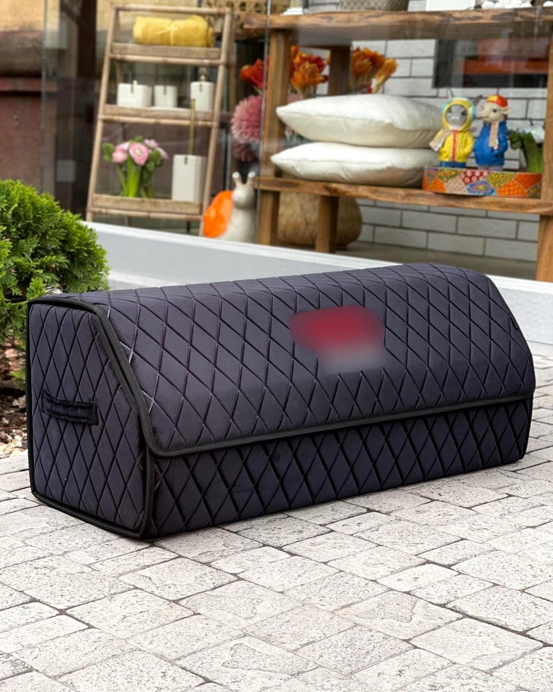 May include: A black, quilted storage container with a diamond pattern. The container has a rectangular shape with a curved top and a handle on the side. The background includes a wooden shelf with decorative items.