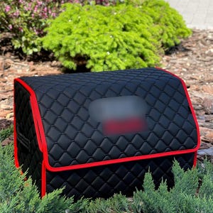 May include: Black quilted car trunk organizer with red trim. The organizer has a rectangular shape with a curved top and a blurred rectangular area. The exterior is a diamond pattern. The organizer is surrounded by green plants and brown mulch.