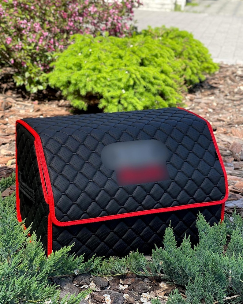 May include: Black quilted car trunk organizer with red trim. The organizer has a rectangular shape with a curved top and a blurred rectangular area. The exterior is a diamond pattern. The organizer is surrounded by green plants and brown mulch.