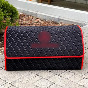 May include: Black, quilted car organizer with red trim. The organizer has a diamond pattern and a rectangular shape. The red trim outlines the top and bottom edges of the organizer. The organizer is designed to store items in a vehicle.