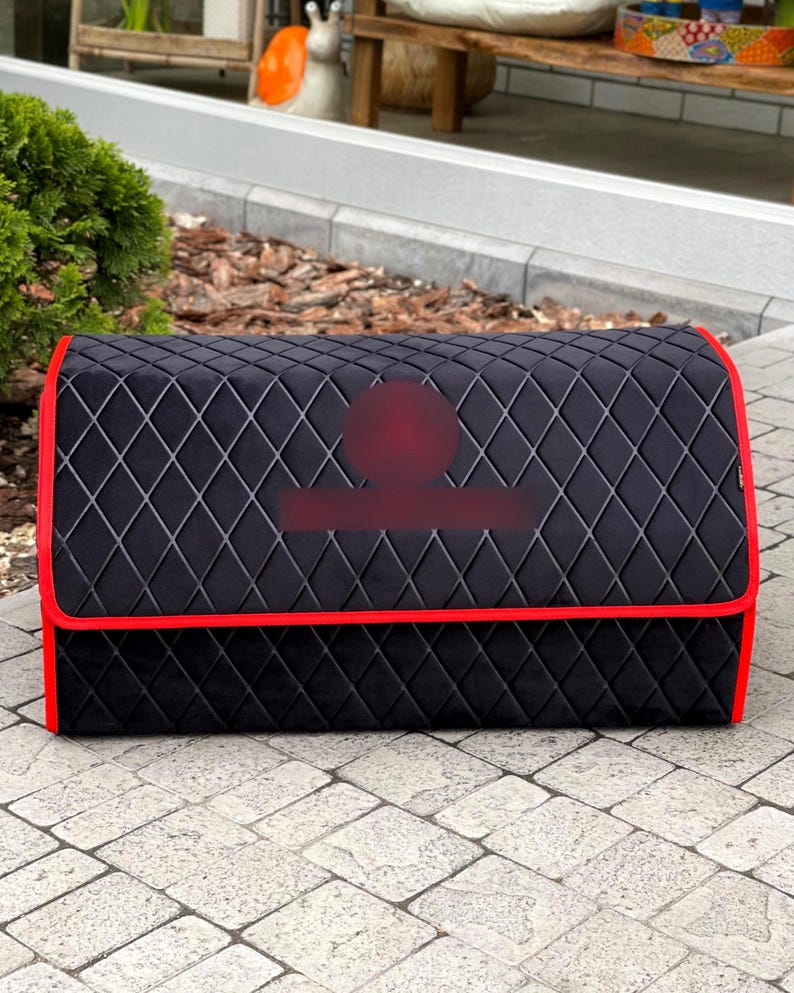 May include: Black, quilted car organizer with red trim. The organizer has a diamond pattern and a rectangular shape. The red trim outlines the top and bottom edges of the organizer. The organizer is designed to store items in a vehicle.