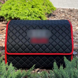 May include: Black quilted car organizer with red trim. The organizer has a diamond pattern and a rectangular shape. The red trim outlines the edges of the organizer. The organizer is designed to store items in a vehicle.