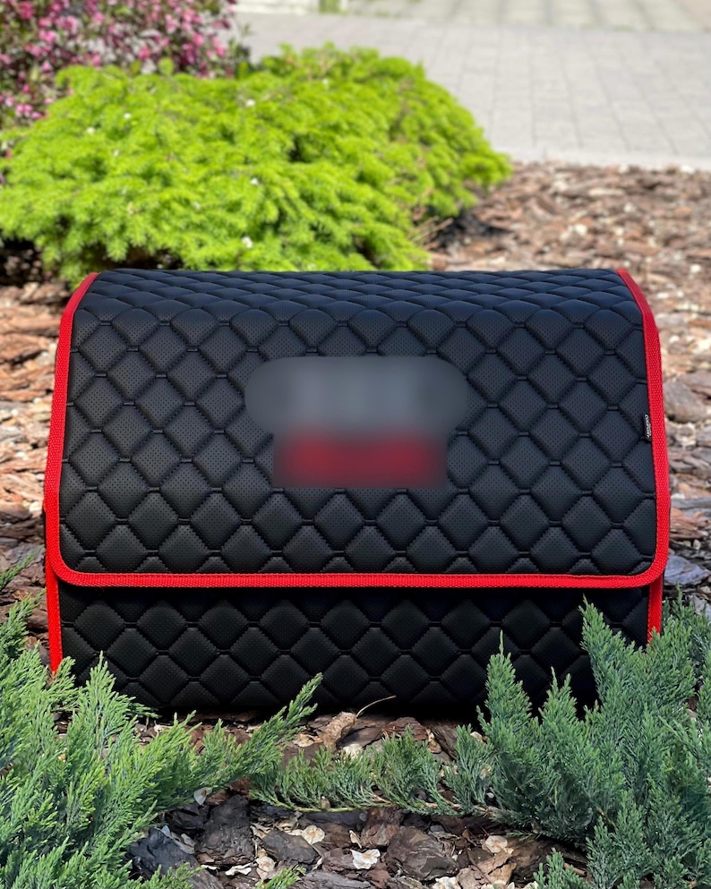 May include: Black quilted car organizer with red trim. The organizer has a diamond pattern and a rectangular shape. The red trim outlines the edges of the organizer. The organizer is designed to store items in a vehicle.