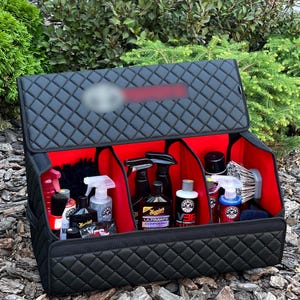 May include: Black quilted car organizer with red interior compartments. The organizer is open, revealing various car detailing products, including spray bottles, waxes, and brushes. The organizer is designed to keep car care items organized.