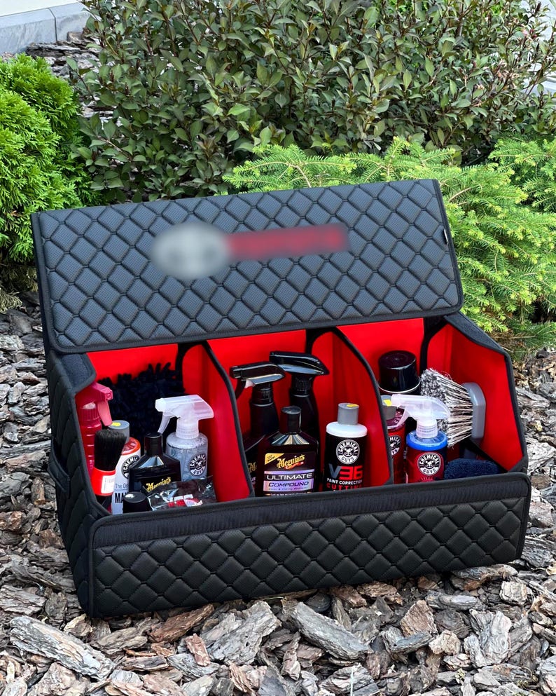 May include: Black quilted car organizer with red interior compartments. The organizer is open, revealing various car detailing products, including spray bottles, waxes, and brushes. The organizer is designed to keep car care items organized.
