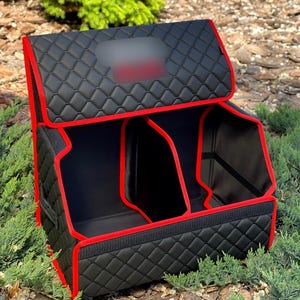 May include: Black car trunk organizer with red trim. The organizer has a quilted pattern and multiple compartments. The lid is open, revealing the interior. The organizer is designed to keep the car trunk tidy.