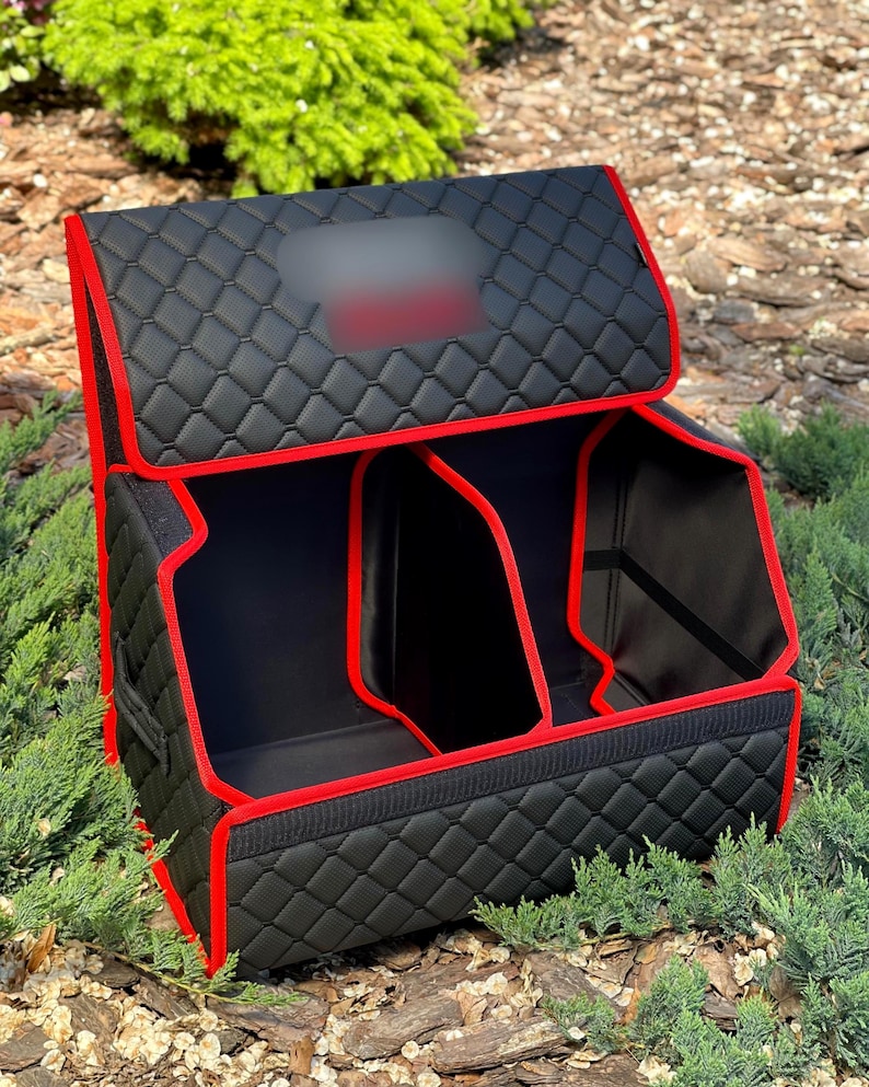 May include: Black car trunk organizer with red trim. The organizer has a quilted pattern and multiple compartments. The lid is open, revealing the interior. The organizer is designed to keep the car trunk tidy.