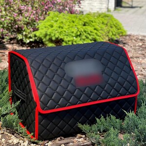 May include: Black, quilted car organizer with red trim. The organizer has a rectangular shape with a hinged top and a blurred rectangular area on the top. The organizer is sitting on a bed of mulch and small green plants.