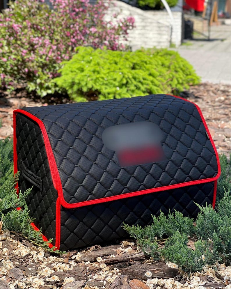 May include: Black, quilted car organizer with red trim. The organizer has a rectangular shape with a hinged top and a blurred rectangular area on the top. The organizer is sitting on a bed of mulch and small green plants.