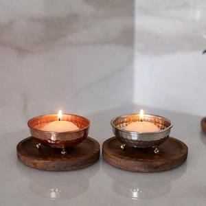 May include: Two lit candles in hammered metal bowls with small brass feet, resting on round wooden coasters. The candles are set against a neutral background, with a vase of flowers in the background.