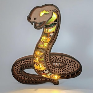 Puede incluir: Una escultura de serpiente de madera con intrincados detalles recortados e iluminación LED cálida. El diseño en capas marrón y beige crea un efecto 3D, con un cuerpo enrollado y la cabeza levantada. La luz brilla a través de los recortes.