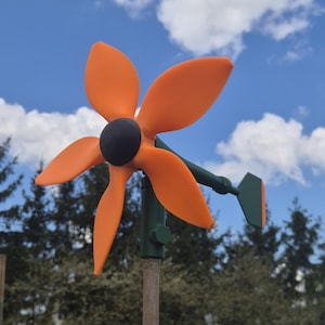 Könnte beinhalten: Ein leuchtend orangefarbenes und schwarzes Windrad mit vier gebogenen Flügeln. Das Windrad ist auf einer grünen Stange und einem Holzpflock montiert. Der Hintergrund zeigt einen blauen Himmel mit weißen Wolken und grünen Bäumen.