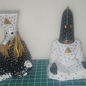 Peut inclure: Deux figurines stylisées en position assise. La figurine de gauche a une tête triangulaire blanche à pois noirs, un long nez et des cheveux dorés. La figurine de droite a une tête conique noire avec des accents dorés et une robe blanche à pois noirs.
