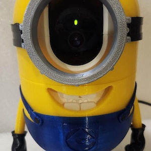 Puede incluir: Una cámara de seguridad amarilla y azul con temática de Minion. La cámara tiene una lente grande y circular y una cara sonriente. Cuenta con un mono azul, guantes y zapatos negros. La cámara está diseñada para parecerse a un personaje de Minion.