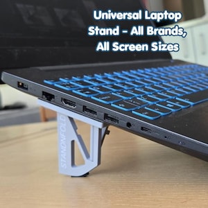 Pode incluir: Um computador portátil com teclado retroiluminado azul é suportado por um suporte dobrável cinzento. O suporte está rotulado "STANDNFOLD" e foi concebido para todas as marcas e tamanhos de ecrã. O texto acima do portátil diz "Universal Laptop Stand - All Brands, All Screen Sizes."