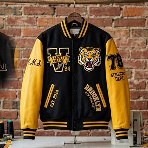 Può includere: Una giacca varsity nera e gialla con una toppa a testa di tigre, la scritta Varsity Champ, Brooklyn New York City e il numero 78. Il retro mostra il profilo dorato di uno skyline con il testo Est. 1978 e il disegno di una scarpa alata.
