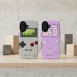 May include: Two phone cases designed to resemble retro handheld game consoles. One is gray with a pixelated game screen, the other is lavender with a cat illustration. Both cases have the text "Your Name".