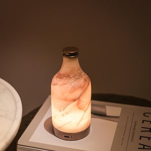May include: A decorative bottle-shaped lamp with a warm, glowing interior. The lamp has a marble-like appearance with shades of peach and white. It features a bronze-colored base and cap, creating a soft, ambient light.