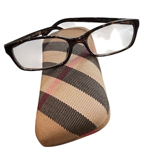 May include: Dark brown eyeglasses with a rectangular frame rest on a beige and black plaid glasses case. The case has a rounded shape and a classic plaid pattern with red accents. The glasses have clear lenses.