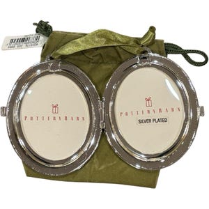 May include: A silver-plated oval locket with a hinged design, featuring two frames for photos. The interior displays the text Pottery Barn and Silver Plated. It is positioned in front of a small olive green fabric drawstring pouch with a ribbon.