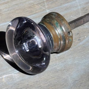 May include: A round, clear glass door knob with a slight purple tint, attached to a weathered, tarnished brass circular base. A dark, square metal spindle extends from the back of the brass mounting, featuring a small screw hole on the side.