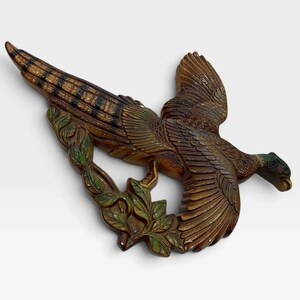 May include: A wall-mounted decorative plaque shaped like a pheasant in flight. The bird features brown and tan feathers with a dark green head and neck. Below the bird, a curved branch with green leaves is integrated into the design.