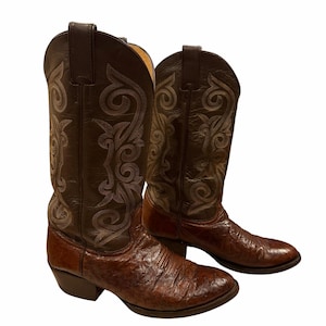 May include: A pair of brown leather cowboy boots featuring a textured, quill-like finish on the foot and a smooth, dark brown shaft adorned with intricate, light-coloured decorative stitching patterns, pull tabs, and a stacked block heel.