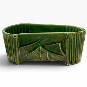 May include: An elongated, oval-shaped ceramic planter with a glossy, deep green glaze. The exterior features a textured design with vertical ribbed columns on the sides and a raised, embossed leaf pattern across the centre front section.