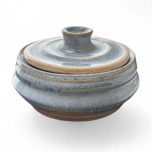 May include: A round ceramic lidded jar featuring a variegated glaze in shades of slate blue, grey, and cream. The vessel has a wide, tapered body with a smooth, circular lid topped by a small, centered knob handle, revealing a brown clay base.
