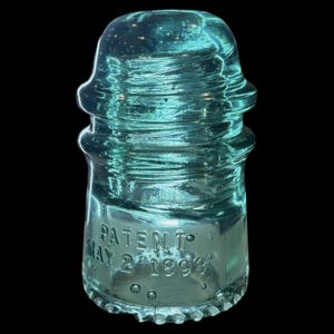 Op de afbeelding: Een doorschijnende aquablauwe glazen elektrische isolator met een afgeronde bovenkant en getrapte ribbels. De basis is voorzien van de in reliëf aangebrachte tekst PATENT MAY 2 1893. Het glas bevat kleine luchtbellen en een glad oppervlak.