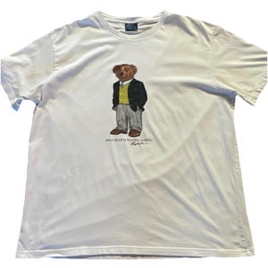 May include: A white short-sleeve t-shirt featuring a graphic of a brown bear wearing a black blazer, yellow sweater vest, white collared shirt, and gray trousers. The text POLO BEAR by RALPH LAUREN is printed in black below the bear graphic.