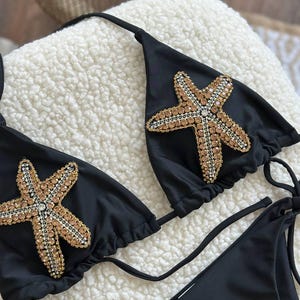 Pode incluir: Um top de biquíni preto com duas grandes aplicações decorativas em forma de estrela-do-mar. Cada estrela é adornada com numerosos cristais pequenos, de cor castanho-claro e transparentes, dispostos num padrão texturado. Possui tiras finas pretas.