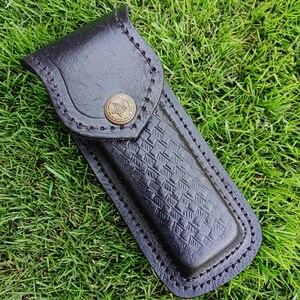 May include: Black leather knife sheath with a decorative button and stitched edges. The sheath has a textured pattern on the front and is designed to hold a knife. The background is green grass.