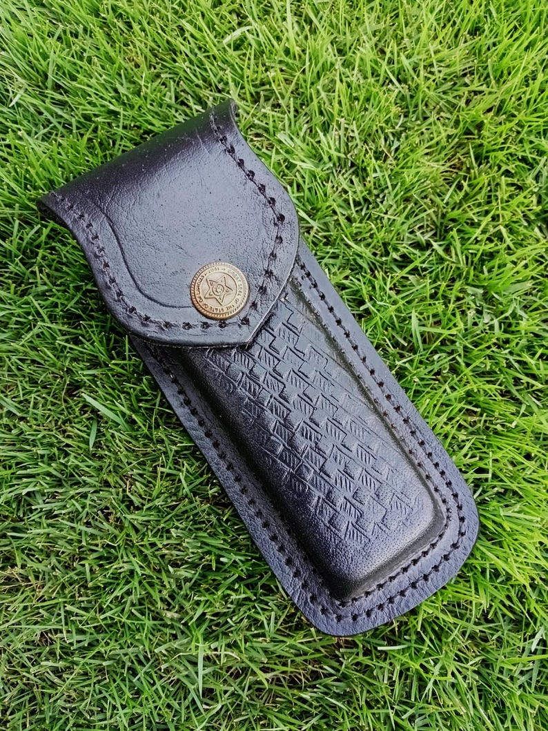 May include: Black leather knife sheath with a decorative button and stitched edges. The sheath has a textured pattern on the front and is designed to hold a knife. The background is green grass.