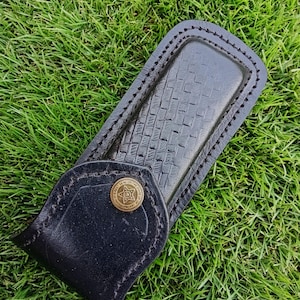 May include: Black leather knife sheath with a basket weave design. The sheath features a stitched border and a flap secured by a brass button with a star emblem. The sheath is on green grass.