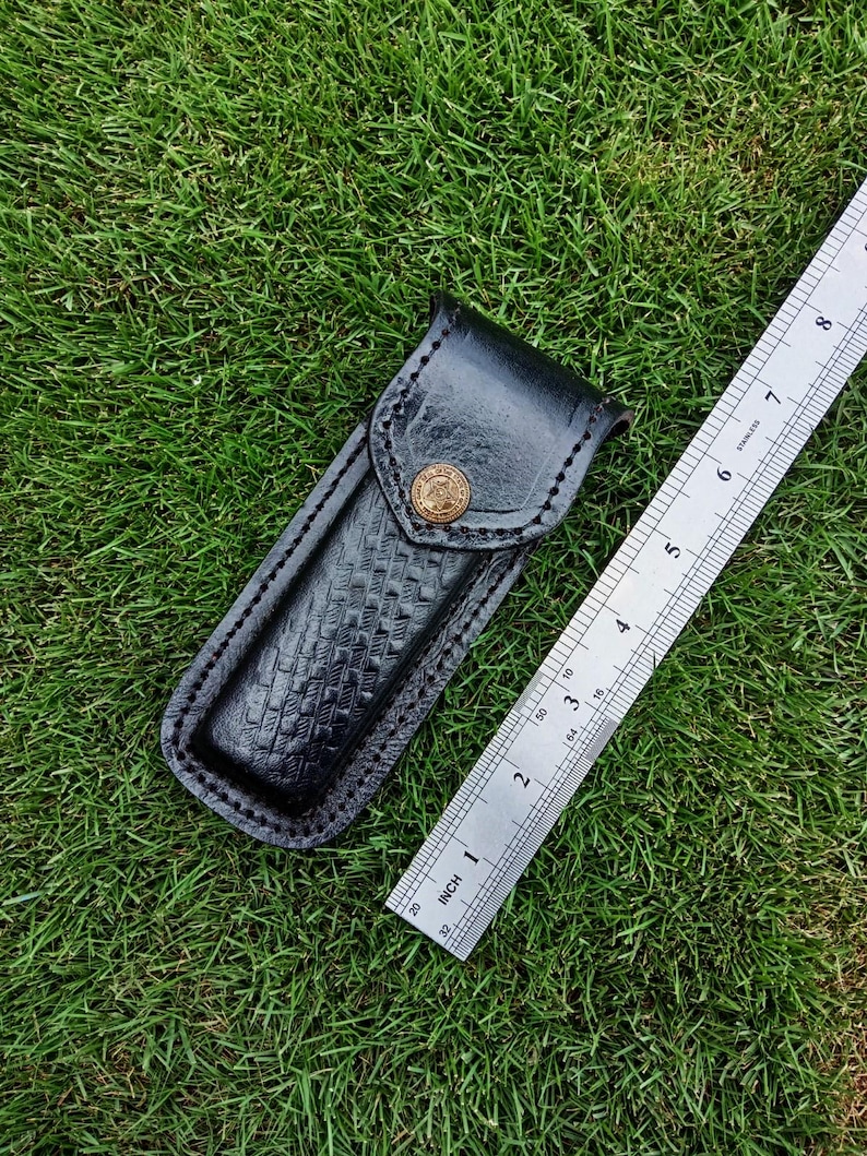 May include: Black leather knife sheath with a basket weave pattern and a brass button. The sheath is next to a stainless steel ruler, showing inches. The sheath is approximately 15 cm long.