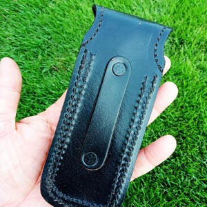 May include: Black leather sheath with a stitched border and a strap closure. The sheath is designed to hold a knife or similar tool. The leather appears smooth and the stitching is a contrasting colour. The sheath is held in a hand against a green background.