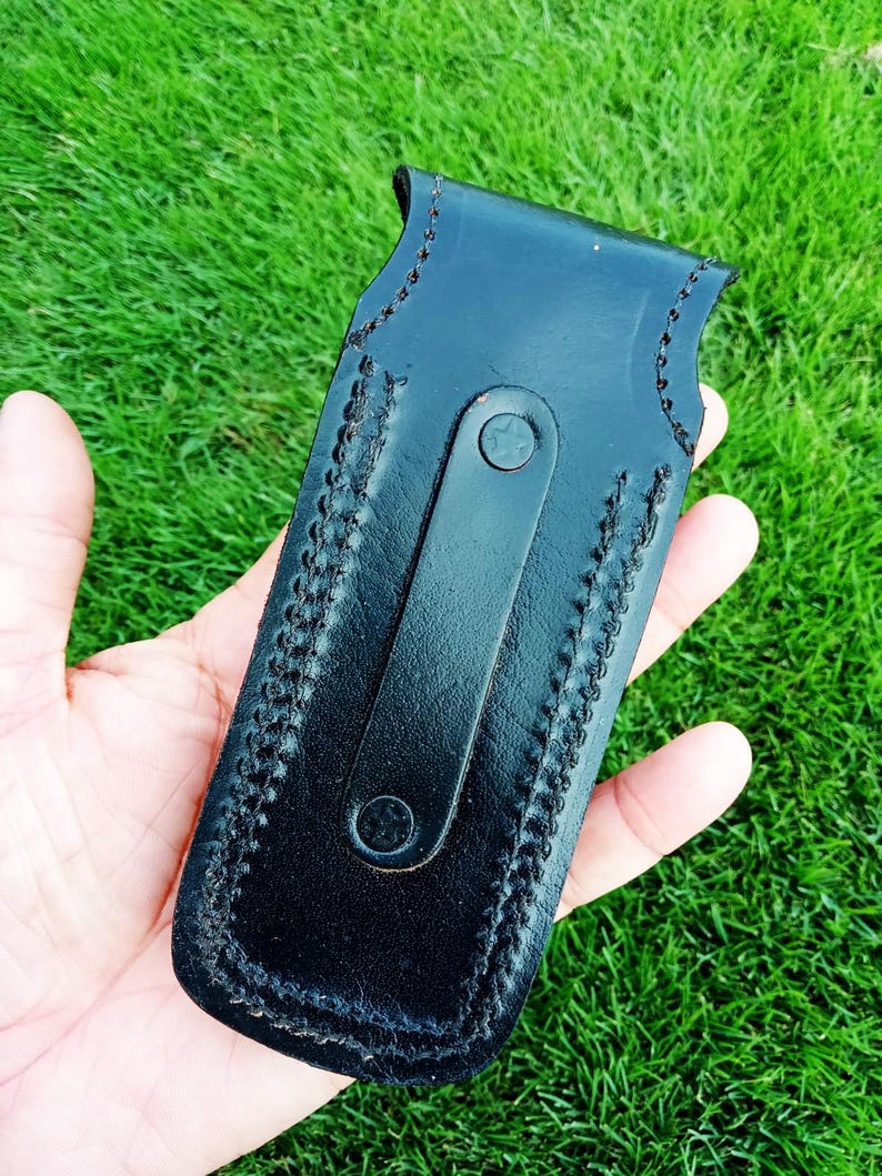 May include: Black leather sheath with a stitched border and a strap closure. The sheath is designed to hold a knife or similar tool. The leather appears smooth and the stitching is a contrasting colour. The sheath is held in a hand against a green background.