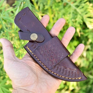 May include: A brown leather knife sheath with decorative stitching and embossed patterns. The sheath has a flap secured by a brass button. The leather is a rich, dark brown color.