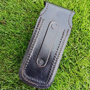 May include: A black leather sheath with a stitched border and a strap closure. The sheath is rectangular with a curved top and is designed to hold a small tool or knife. The leather has a slightly glossy finish.