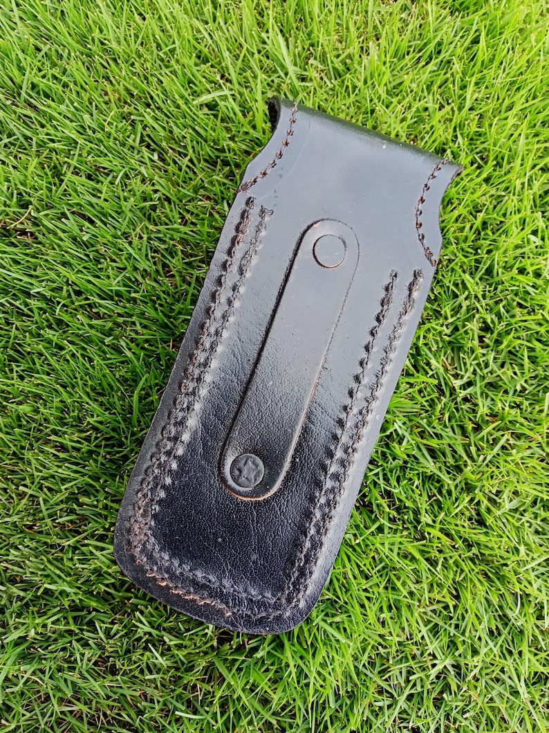 May include: A black leather sheath with a stitched border and a strap closure. The sheath is rectangular with a curved top and is designed to hold a small tool or knife. The leather has a slightly glossy finish.