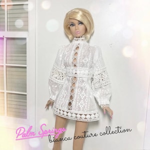 May include: A fashion doll with blonde hair wearing a white lace mini dress featuring long sleeves, a high neckline, and a vertical cutout pattern down the centre. The text Palm Springs bianca couture collection is printed in pink and black.